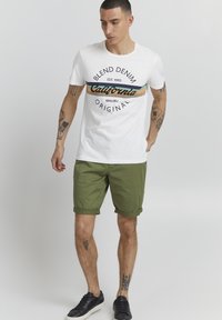 Hvit t-skjorte med et flerfarget "California" grafikk, kombinert med grønne shorts. Svarte joggesko fullfører antrekket.