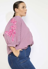Blusa a quadri rosa e bianchi con maniche a palloncino, caratterizzata da un vivace ricamo floreale rosa sulle spalle. Abbinata a jeans blu.