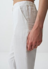 Pantalons en lin blanc avec une coupe décontractée, dotés d'une taille élastique et de poches latérales. Texture lisse avec un design minimaliste.