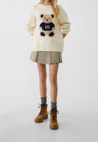 Kvinna i krämfärgad tröja med teddygrinddesign, rutmönstrad plisserad kjol, grå strumpor och beige snörstövlar som står på vit bakgrund.