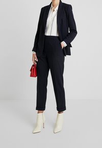 Costume sur mesure bleu marine à pois avec un blazer cintré et un pantalon retroussé. Chemise blanche à boutons avec des rayures. Bottines crème. Sac à main rouge.