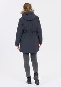 Dunkelblaue Parka mit gefütterter Kapuze, tailliertem Schnitt und Rückenschlitzen, kombiniert mit grauen Leggings und schwarzen Stiefeln.