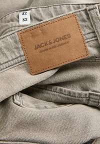 Pilkos džinsinės kelnės su rudono odos etiketė, ant kurios užrašyta "JACK & JONES JEANS INTELLIGENCE." Dydžių etiketės nurodo 32 colių matavimus.
