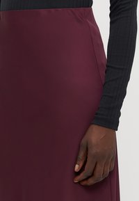 Gonna bordeaux a vita bassa in tessuto morbido con design aderente. Esposta con un top nero a maniche lunghe a coste e una mano appoggiata sulla gonna.