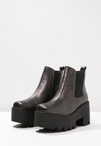 Botas tobilleras de color gris metálico con suelas de plataforma negra gruesa, paneles laterales elásticos y una textura suave. Diseño minimalista con puntera redondeada.