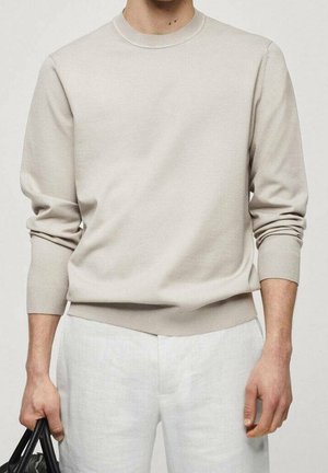 Mand iført beige langærmet sweater og hvide bukser, holder en sort taske i venstre hånd mod en neutral baggrund.
