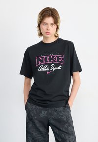 Black cotton t-shirt with a bold pink and white "NIKE Athletic Depart." graphic. Short sleeves and a relaxed fit. Textured black patterned pants.