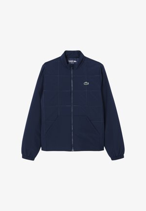 Giacca blu navy con cerniera, cuciture a griglia, polsini elastici, due tasche frontali e un piccolo logo a coccodrillo verde sul petto.