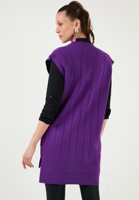 Purple knitted sleeveless cardigan with vertical ribbed texture, side slits, and a relaxed fit, worn over a black long-sleeve shirt.