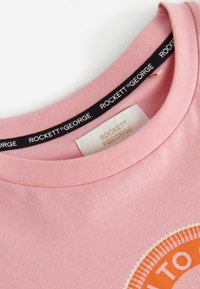 Camiseta rosa de cuello redondo con etiqueta "Rockett St George" en el interior del cuello y gráfica circular naranja parcialmente visible en el frontal.