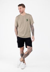 Hombre con tatuajes en brazos y piernas que lleva una camiseta beige con diseño de palmera, pantalones cortos negros y zapatillas blancas, de pie sobre un fondo blanco.