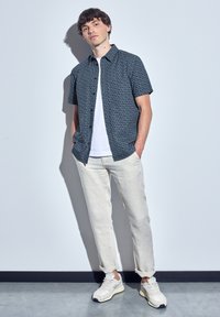 Short-sleeve, dark patterned shirt over a white T-shirt, paired with light beige trousers and gray sneakers, standing against a neutral background.