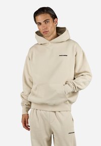 Beige hettegenser med stor frontlomme, snor i hetta, og liten svart "PEGAJOR" logo på brystet. Laget av mykt bomullsmateriale.