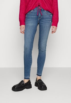Femme portant un jean skinny bleu avec des fermetures éclair à la cheville, des mocassins à plateforme noirs, et un pull rose vif avec un cordon de serrage à l'avant.