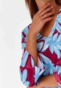 Kukallinen mekko, jossa on marooni tausta ja suuria sinisiä kukkia, V-kaula-aukko ja kolme neljäsosaa hihat. Pehmeä kangasrakennetta näkyvissä.