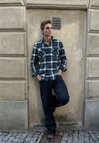 Ung man i blå och grön rutig skjorta och mörka jeans lutar mot en beige vägg med händerna i fickorna, stående på kullerstensbeläggning.