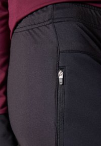 Svart treningsbukser med glidelåslomme på siden, elastisk midje og glatt, stretchy stoff. Overdelen er mørkerød.