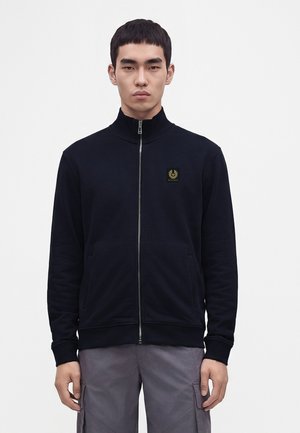 Junger Mann trägt eine schwarze Jacke mit Reißverschluss und einem Logo auf der linken Brustseite sowie graue Cargohosen, steht vor einem schlichten weißen Hintergrund.