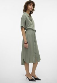 Robe boutonnière vert sauge avec une ceinture, manches courtes, fentes latérales et longueur midi, portée avec des escarpins noirs à bout pointu. Texture lisse.