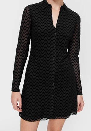 Zwarte jurk met lange mouwen en knopen, met een golvend doorschijnend patroon en kraag, gedragen tot halverwege het bovenbeen.