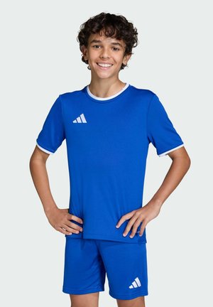 Uśmiechający się chłopiec z kręconymi włosami, ubrany w niebieską sportową koszulkę Adidas i pasujące do niej spodenki, z rękami na biodrach, zwrócony przodem.