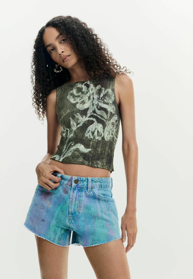 Femme aux cheveux bouclés portant un haut sans manches vert à motifs floraux et un short en jean bleu tie-dye, debout devant un fond clair uni.