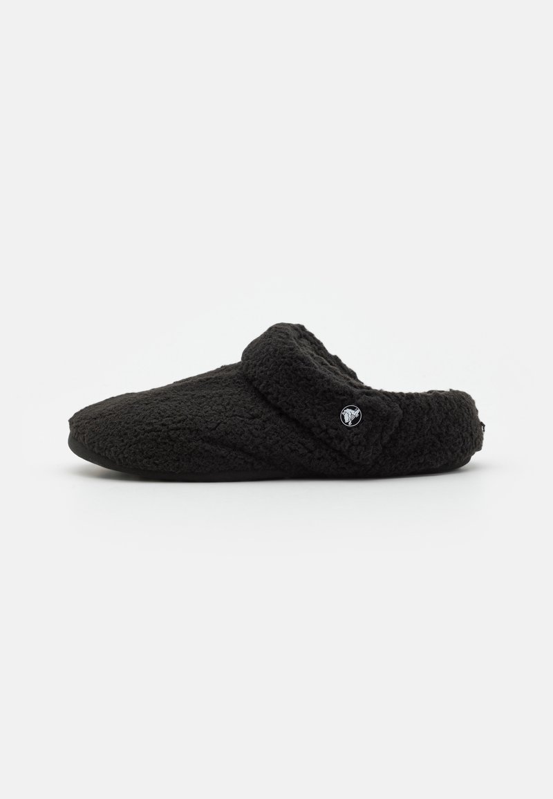 Chaussure noire souple texturée à enfiler avec une large bride au talon et un petit logo rond sur le côté, présentée sur un fond blanc uni.