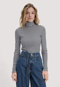 Gestreiftes Rollkragenoberteil in Schwarz und Weiß, taillierte Schnittform mit langen Ärmeln. Getragen mit hoch taillierten blauen Jeans, die einen Bindegürtel besitzen.