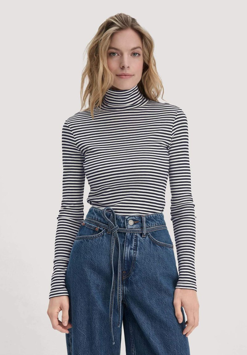 Gestreiftes Rollkragenoberteil in Schwarz und Weiß, taillierte Schnittform mit langen Ärmeln. Getragen mit hoch taillierten blauen Jeans, die einen Bindegürtel besitzen.