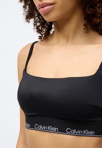 Μαύρο αδιάκοπο σουτιέν με λεπτές τιράντες, με φαρδιά ελαστική ταινία με το λογότυπο "Calvin Klein" σε λευκό χρώμα. Ομαλή υφή υφάσματος.