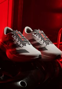 Weiße Sportschuhe mit schwarzen Streifen und grauen Schnürsenkeln, von rotem Licht beleuchtet, ruhen auf schwarzem metallischem Fitnessgerät.