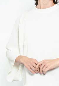 Blusa bianca in tessuto leggero, con colletto alto, dettagli plissettati sulle spalle e maniche fluenti. Texture morbida e design pulito.