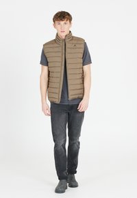 Bruine gequiltte vest met een hoge kraag, gedragen over een grijze shirt. Het model draagt ook een donkere spijkerbroek en grijze laarzen. Eenvoudig, functioneel ontwerp.