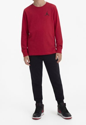 Barn iført rød langærmet skjorte med sort logo, sorte bukser og sorte og røde høje sneakers, stående mod hvid baggrund.