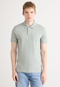 Ung mann iført en lys grønn polo-skjorte og blå jeans, står mot en ensfarget hvit bakgrunn, vendt mot kameraet med et nøytralt uttrykk.