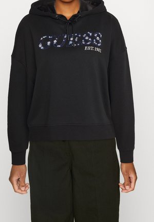 Sudadera negra Guess con logo de lentejuelas y texto "EST. 1981" llevada por persona con pantalones verde oscuro, manos relajadas a los lados.