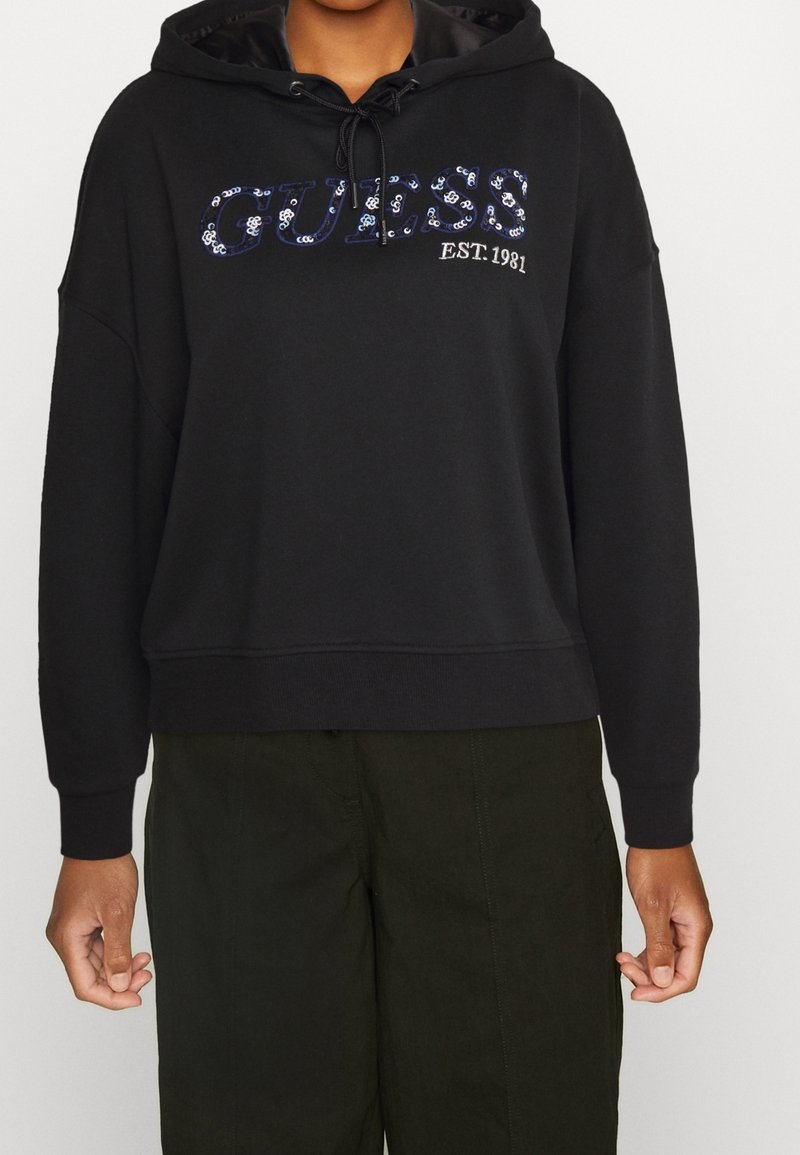 Sweat à capuche noir Guess avec logo en sequins et texte "EST. 1981" porté par une personne avec un pantalon vert foncé, mains détendues le long du corps.