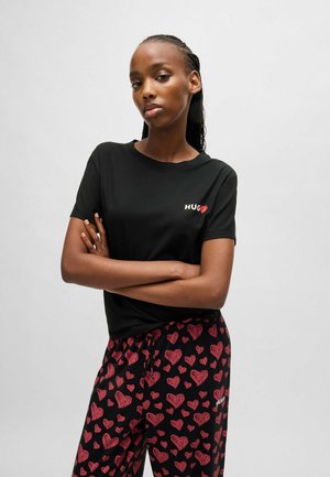 Black cotton t-shirt with "HUG" logo in white and red. Paired with black pants featuring red heart prints. Model stands with arms crossed.
