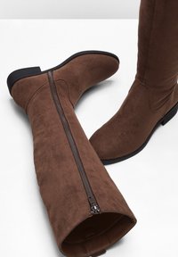 Stivali alti fino al ginocchio in suede marrone con cerniera laterale, suola piatta e punta leggermente appuntita. Texture liscia e design minimalista.