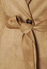 Beżowa zamszowa kurtka o teksturowanej powierzchni, z wiązanym paskiem w talii i dużym okrągłym guzikiem z przodu.