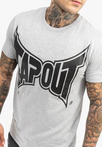 Pelēka kokvilnas T-kreklā ar lielu melnu "TAPOUT" logo uz priekšpuses, īsām piedurknēm un apaļu kakla izgriezumu.