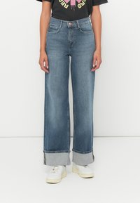 Široke denim hlače svetlo modre barve s prepognjenim sivim robom. Vključujejo tradicionalne žepe in zaponko na gumb. Združene z belimi supergami.