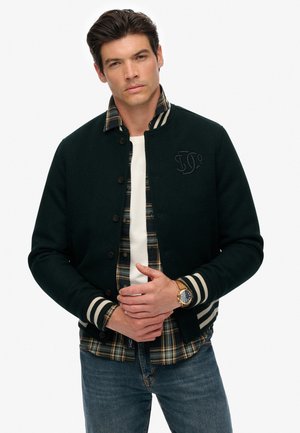 Mørkegrøn uldjakke med stribede manchetter, knappet front og broderet logo, lagdelt over en ternet flannel skjorte. Klassiske denim jeans.