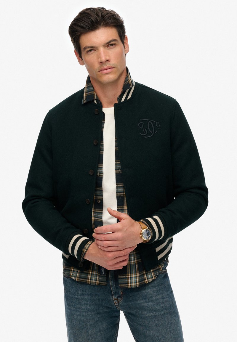 Mørkegrøn uldjakke med stribede manchetter, knappet front og broderet logo, lagdelt over en ternet flannel skjorte. Klassiske denim jeans.