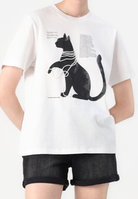T-shirt in cotone bianco con una silhouette nera di un gatto decorata con perline simili a perle, accompagnata da un testo stampato accanto al design.