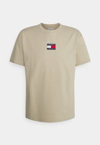 Beige short-sleeve T-shirt with crew neck, featuring a small Tommy Jeans logo patch in navy, white, and red on the chest.