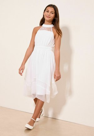 Noor tüdruk kannab valget varrukateta pitsiliste detailidega kleiti ja kõnnib enesekindlalt valgetes pahkluu rihmaga kingades ühtlase beeži taustaga.