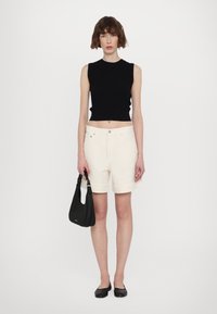 Svart ribbet ermeløs topp, kremfargede shorts med oppbrett og svart håndveske. Modellen har på seg svarte flate sko. Enkelt, tettsittende design.