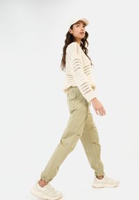 Gestrickter cremefarbener Pullover mit offener Webart, olivgrüne Cargo-Shorts und helle Sneakers. Hat eine lockere Passform und gerippte Bündchen. Wirkt lässig und bequem.