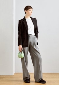 Svart blazer över en vit skjorta, grått rutigt byxor och svarta skor. Håller en liten grön handväska. Texturerat trägolv.